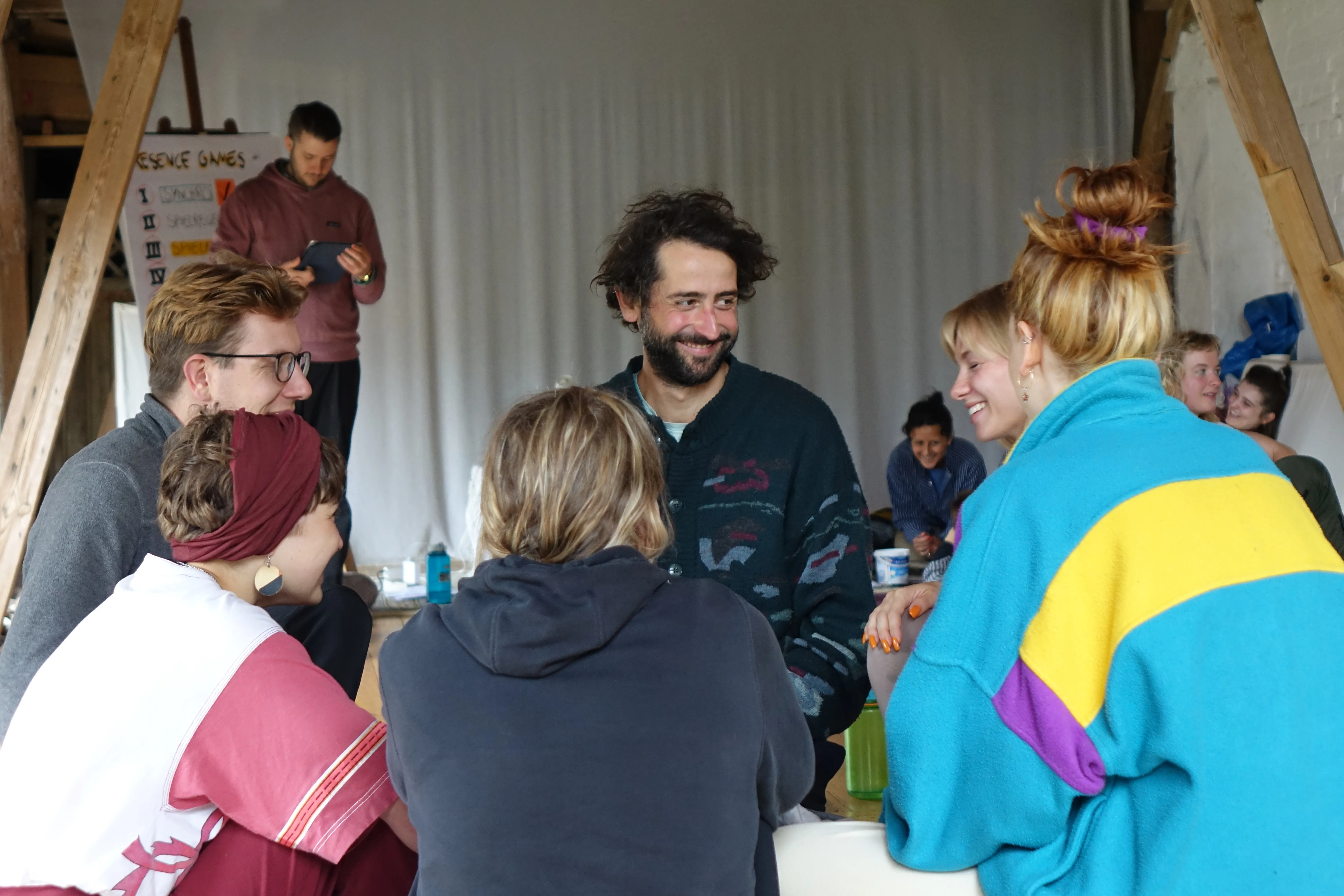 Menschen sitzen in einem Kreis und unterhalten sich. Der Mann im Fokus lächelt die Frau neben sich an. Im Hintergrund sind andere Workshop-Teilnehmende zu sehen.